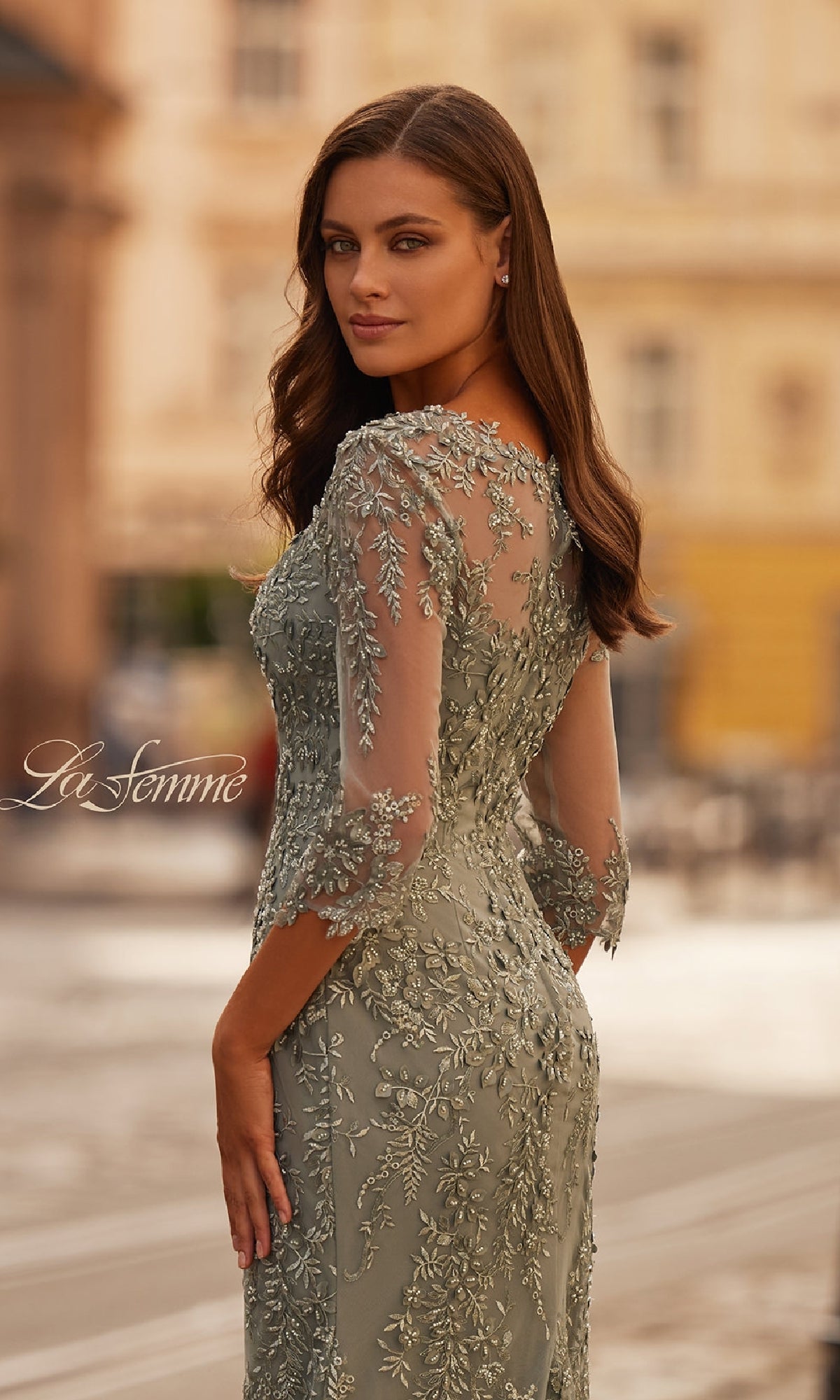 A woman in the La Femme 31909 long formal dress, featuring light green lace, three-quarter sleeves, and sheer details, stands outdoors looking over her shoulder against an elegant city backdrop.